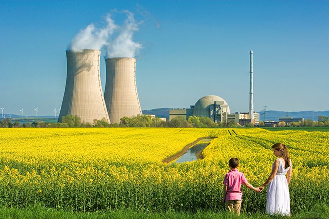 Rear view on Young mother holding hand of her son looking concerned at him in front of flowering canola field and nuclear power station. Some small wind turbines in the background on the left side of the two cooling towers. Useful as symbol for future and energy questions.